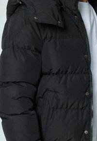 Doudoune noire à conception matelassée, fermeture éclair, boutons-pression et poche latérale. Le tissu présente une texture lisse et douce.