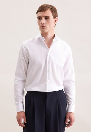 Jonge man met kort bruin haar, gekleed in een wit overhemd met knopen en donkere geplooide broek tegen een lichte neutrale achtergrond.