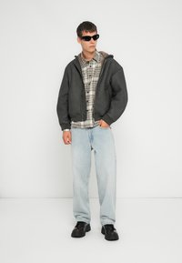 Veste noire courte avec capuche sur une chemise à carreaux, jeans bleu clair et chaussures noires. Modèle portant des lunettes de soleil sombres, se tenant devant un fond blanc.