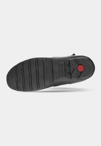 Semelle de chaussure en caoutchouc noir avec un motif de grip texturé, des rainures horizontales et un accent circulaire rouge distinctif sur le talon.