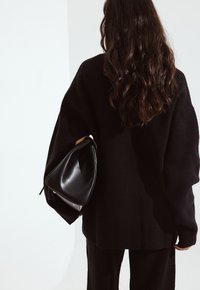 Črna prevelika jopica z ribastimi manšetami, v kombinaciji z gladko črno torbico za ramo, ki ima zaobljeno obliko in subtilne kovinske detajle.