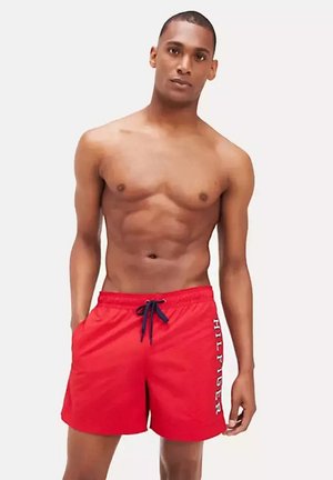 Man draagt rode zwemshorts met de tekst "HILFIGER", staat ontspannen tegen een effen lichte achtergrond.