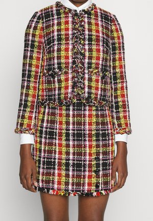 Femme portant une veste et une jupe en tweed à carreaux rouge, jaune, noir et blanc, avec des bords effilochés et des détails de boutons.