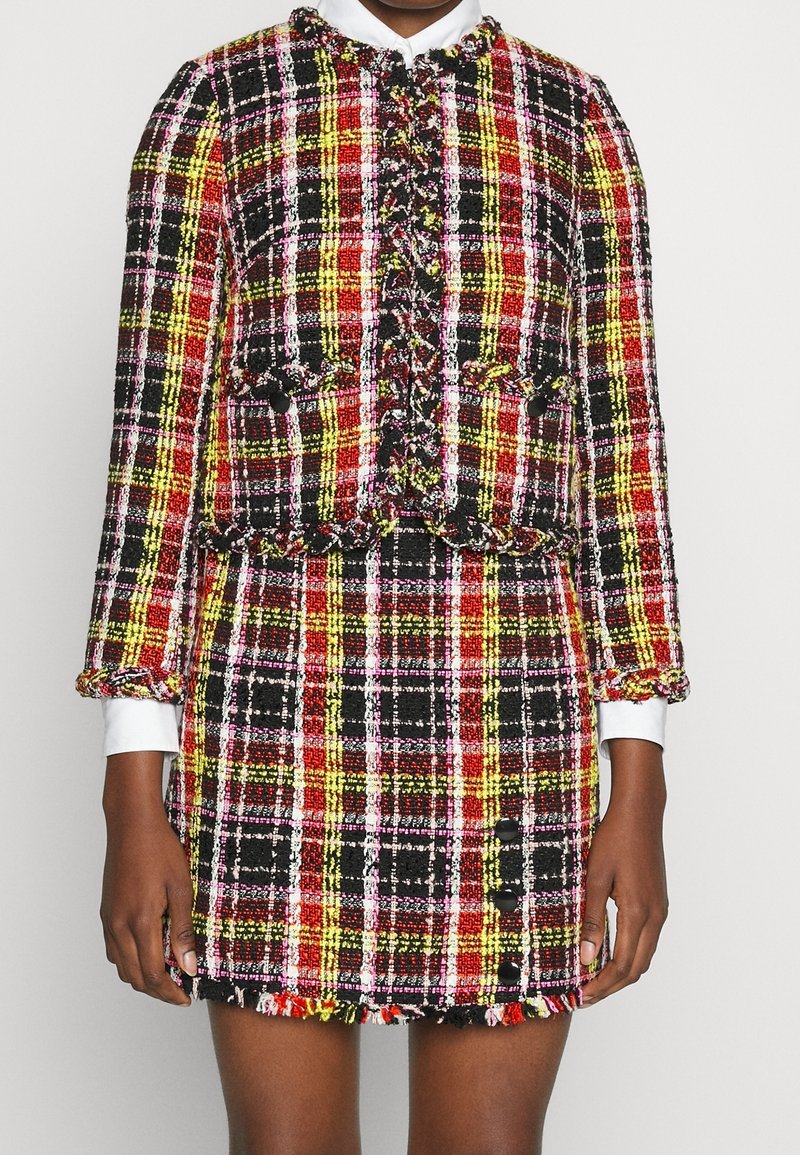 Femme portant une veste et une jupe en tweed à carreaux rouge, jaune, noir et blanc, avec des bords effilochés et des détails de boutons.