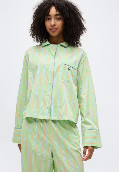 Νεαρή γυναίκα με σγουρά μαλλιά φοράει ελαφρύ πράσινο ριγέ πιτζάμα με κουμπιά, μακριά μανίκια και ταιριαστό παντελόνι, σε λευκό φόντο.