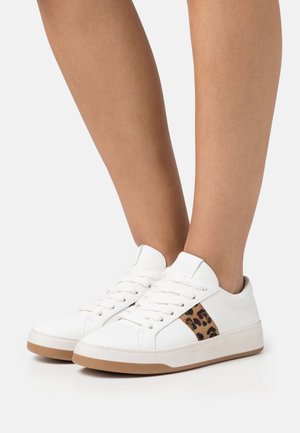 Jambes portant des baskets basses blanches avec un détail imprimé léopard sur le côté et des semelles marron clair sur un fond uni.