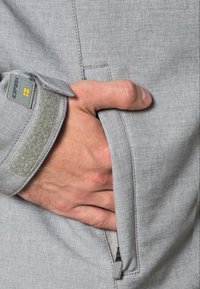 Main masculine à la peau claire insérée dans la poche zippée avant d'une veste gris clair avec une bande Velcro au poignet.