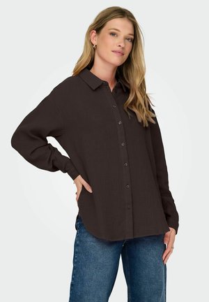 Hnedá košela na gombíky vyrobená z textúrovanej látky, s golierom, dlhými rukávmi a zakriveným spodným lemom. V kombinácii s modrými džínsami.