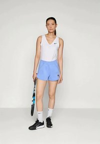 Λευκή αμάνικη μπλούζα με μαύρο λογότυπο Nike, μπλε αθλητικά σορτς, μαύρα παπούτσια και λευκές κάλτσες, κρατώντας ένα ρακέτα τένις.