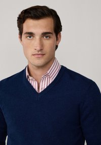 Suéter azul marino de cuello en V sobre una camisa con cuello a rayas en rosa claro y blanco, creando un look superpuesto con texturas y colores distintivos.