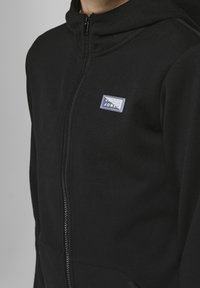 Svart zip-hoodie med en liten rektangulär lapp som står "JACK & JONES" på vänster bröst, bärs av en person.