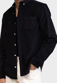 Hombre con una camisa de pana negra abotonada sobre una camiseta negra y pantalones blancos, con una mano en el bolsillo, frente a un fondo liso.