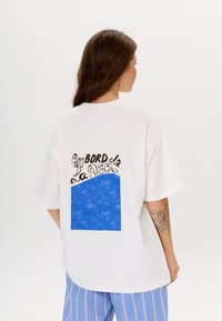 Witte oversized t-shirt met een blauw en crème graphic op de achterkant met de tekst "Au Bord de La Mer" en een gestructureerd blauw vierkant.