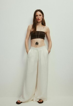 Žena stojící s rukama v kapsách, nosí černý top bez ramínek, bílé široké kalhoty, černé sandály a dlouhý přívěškový náhrdelník.