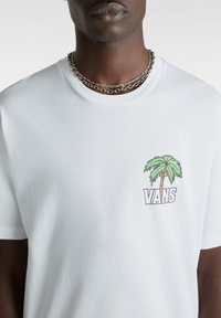 Hvid kortærmet T-shirt lavet af bomuld. Har et grønt palmetræ og "VANS"-logoet trykt på venstre brystområde. Enkel design.