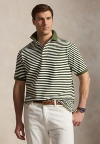 Homme portant un polo à rayures vertes et blanches, un pantalon blanc, une ceinture tressée marron et une montre-bracelet, debout devant un fond neutre.