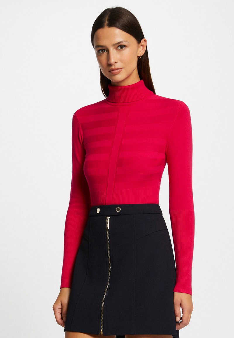 Suéter ajustado de manga larga con cuello alto en un brillante color rosa, con textura acanalada y rayas horizontales, combinado con una falda negra con cremallera.