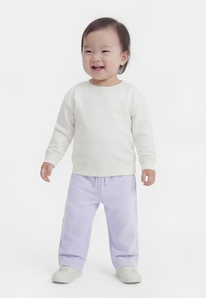 Tout-petit debout et souriant, portant un sweat-shirt blanc, un pantalon violet clair et des chaussures blanches sur un fond blanc.