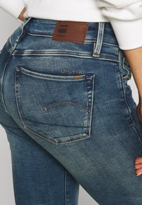 Rå denimjeans i mörkblått med en lätt blekning. Märkbar brun läderpatch i midjan och en böjd stickad bakfickdesign.