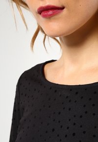 Czarna bluzka z okrągłym dekoltem, krótkimi rękawami i fakturowaną powierzchnią z wypukłymi kropkami. Gładki materiał o miękkim wykończeniu.