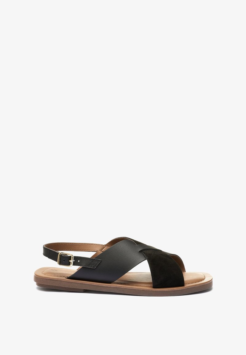 Sandale en cuir noir et daim avec brides croisées, boucle réglable à la cheville et semelle plate. Texture lisse avec un design minimaliste.
