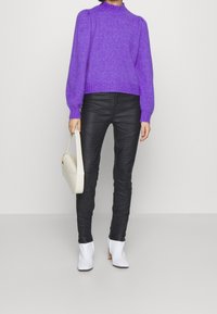 Suéter de punto morado con cuello alto, combinado con pantalones de cuero negros con detalles acolchados y botines blancos hasta el tobillo. Sosteniendo un bolso de mano crema.