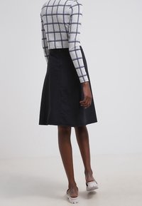 Jupe noire en ligne A avec une texture lisse, longueur genoux, avec un subtil détail de couture. Portée avec un haut à manches longues à carreaux gris.