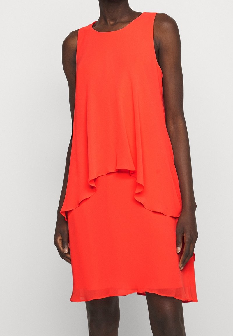 Ärmlös ljusröd lager-på-lager klänning knä-lång som bärs av en stående person framför en ljus enfärgad bakgrund.