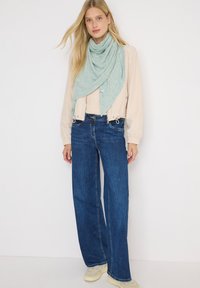 Donna bionda che indossa una giacca beige chiaro, una sciarpa verde chiaro, jeans a gamba larga blu scuro e sneakers avorio in piedi contro uno sfondo bianco.