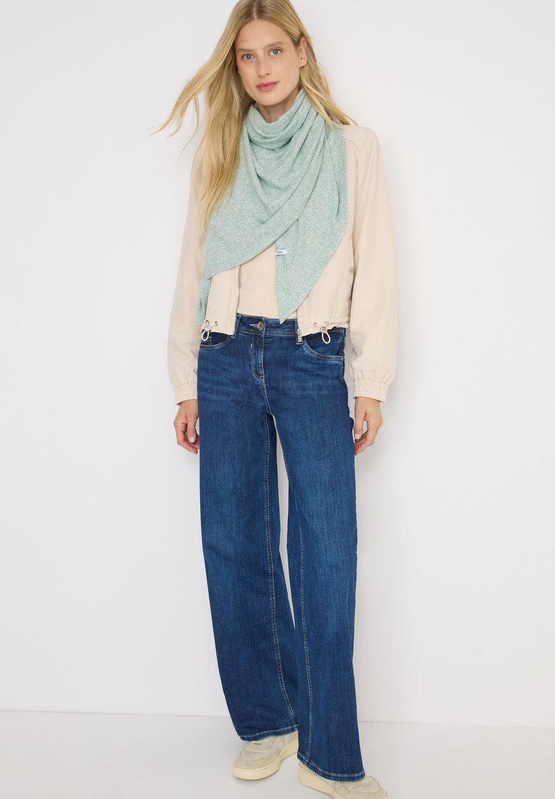 Blonde vrouw in een lichtbeige jas, lichtgroene sjaal, donkerblauwe wijde jeans en off-white sneakers, staand tegen een witte achtergrond.