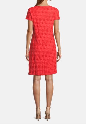 Vrouw staand met de rug naar voren, gekleed in een textuurrode jurk tot op de knie en beige sandalen met hoge hakken, tegen een effen achtergrond.