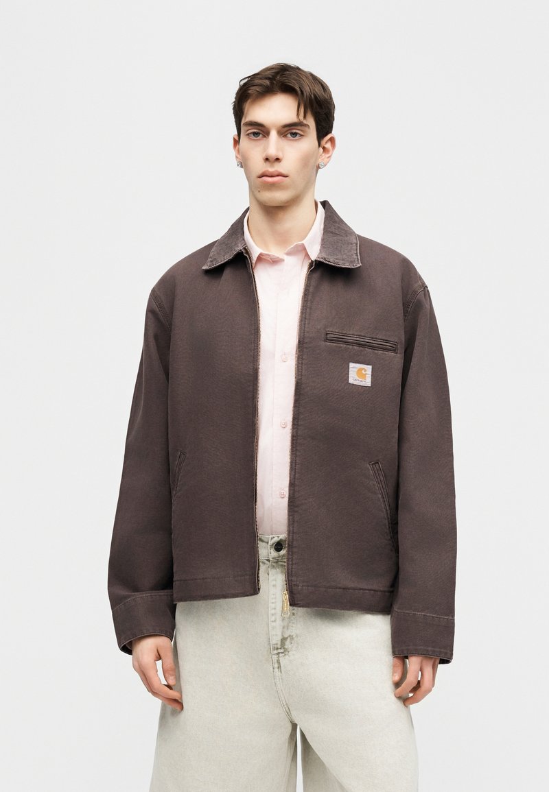 Joven con chaqueta marrón con cremallera sobre una camiseta rosa pálido y pantalones claros, de pie frente a un fondo claro y liso.