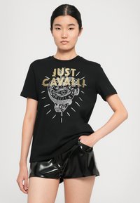 Young woman with black hair wears black T-shirt with gold "JUST CAVALLI" text and white snake and eye graphic, paired with black shiny shorts.