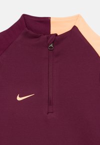 Burgundowy sportowy sweter z pół-zamkiem, kontrastującym brzoskwiniowym panelem na ramionach i małym beżowym logo Nike na klatce piersiowej. Gładki materiał.