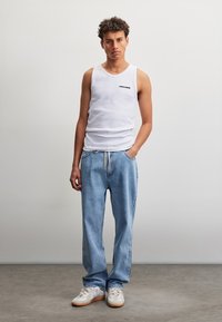Jeune homme debout devant un fond uni, portant un débardeur blanc, un jean bleu clair et des baskets blanches, la main gauche dans la poche.