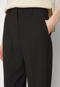 Černé kalhoty na míru z hladkého materiálu, které mají boční kapsy, plochý přední díl a úzký střih. Pas je decentní s poutky na pásek.