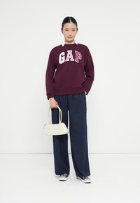 Bordeaux sweatshirt met "GAP" tekst, witte strepen bij de hals, gecombineerd met marineblauwe streepjesshort met wijde pijpen. Witte handtas en sneakers.