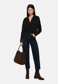 Schwarze Wickelbluse mit langen Ärmeln, dunkelblaue weite Jeans und braune Wildleder-Stiefeletten mit Absatz. Trägt eine große braune Wildleder-Handtasche.
