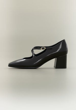 Chaussure Mary Jane en cuir verni noir à bout pointu, talon bloc et fine bride avec boucle dorée sur le côté.