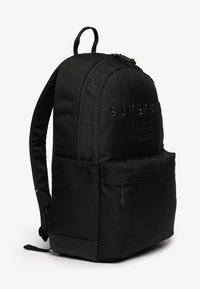 Mochila de tela negra con logo de marca en relieve, compartimento principal con cremallera, bolsillo frontal, correas ajustables y base reforzada.