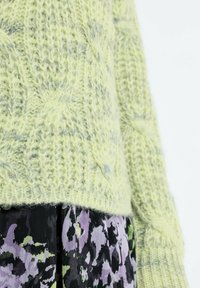 Jasnozielony, gruby sweter z warkoczowym wzorem nałożony na spódnicę w czarno-fioletowo-zielone kwiatowe wzory, pokazujący szczegóły środkowej części tułowia i ramienia.