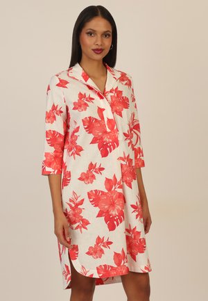 Woman wearing knee-length white dress with red floral print and three-quarter sleeves, standing against plain background.