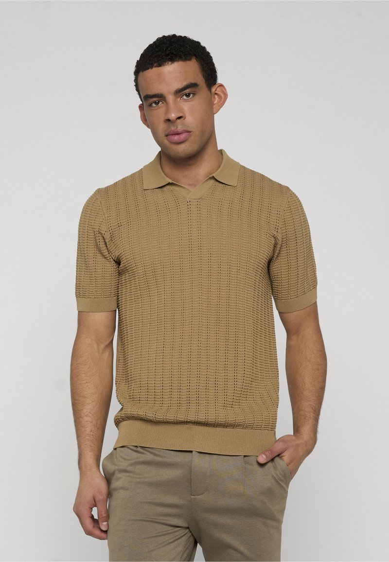 Homme portant un polo en maille beige texturé à manches courtes et un pantalon assorti, debout avec une main dans la poche contre un fond uni.