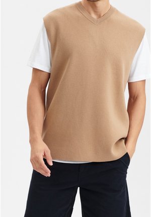 Gilet en tricot à manches courtes de couleur brun clair avec un décolleté en V, porté sur un t-shirt blanc, associé à un short foncé. Texture douce, coupe décontractée.