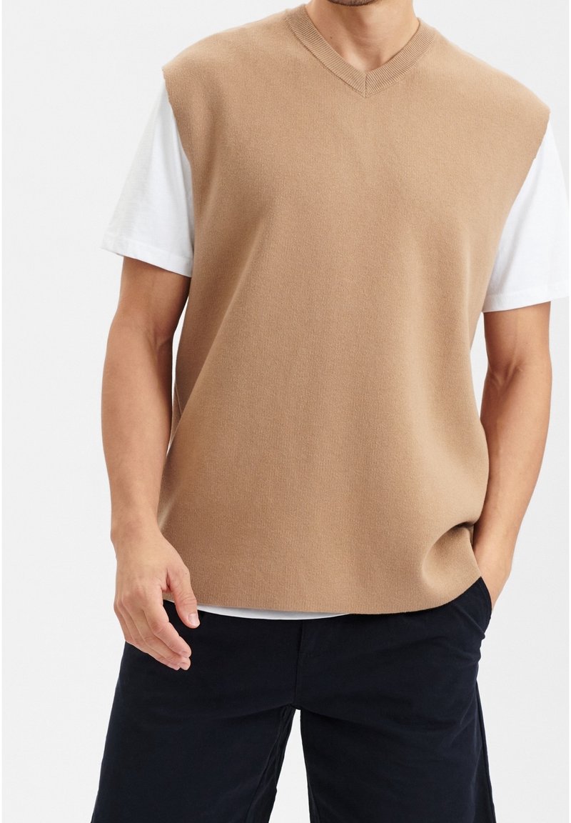 Ärmelloser, hellbrauner Strickpullover mit V-Ausschnitt, getragen über einem weißen T-Shirt und kombiniert mit dunklen Shorts. Weicher Stoff, lässige Passform.