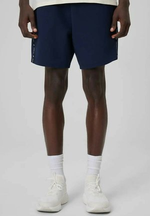 Osoba nosząca granatowe krótkie spodenki sportowe, białe skarpety i białe buty do biegania, stojąca na jednolitym tle.