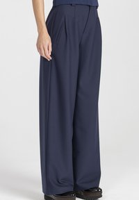 Granatowe spodnie o szerokich nogawkach z przodu z plisami, z wysokim stanem i bocznymi kieszeniami, wykonane z gładkiej tkaniny. Charakteryzują się czystą, dopasowaną sylwetką.