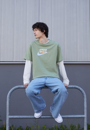 T-shirt verde in cotone con logo Nike colorato, sovrapposto a una maglia bianca a maniche lunghe. Jeans in denim a gamba larga di colore azzurro chiaro e sneakers bianche.