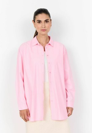 Woman with dark hair tied back wearing gold earrings, a loose light pink button-up shirt over a white top and cream skirt, standing against white background.