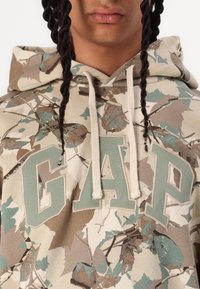 Hoodie med kamouflage-mönster i beige, brunt och teal. Har en stor "GAP"-logotyp i dämpat grönt, med snoddar och en mjuk textur.